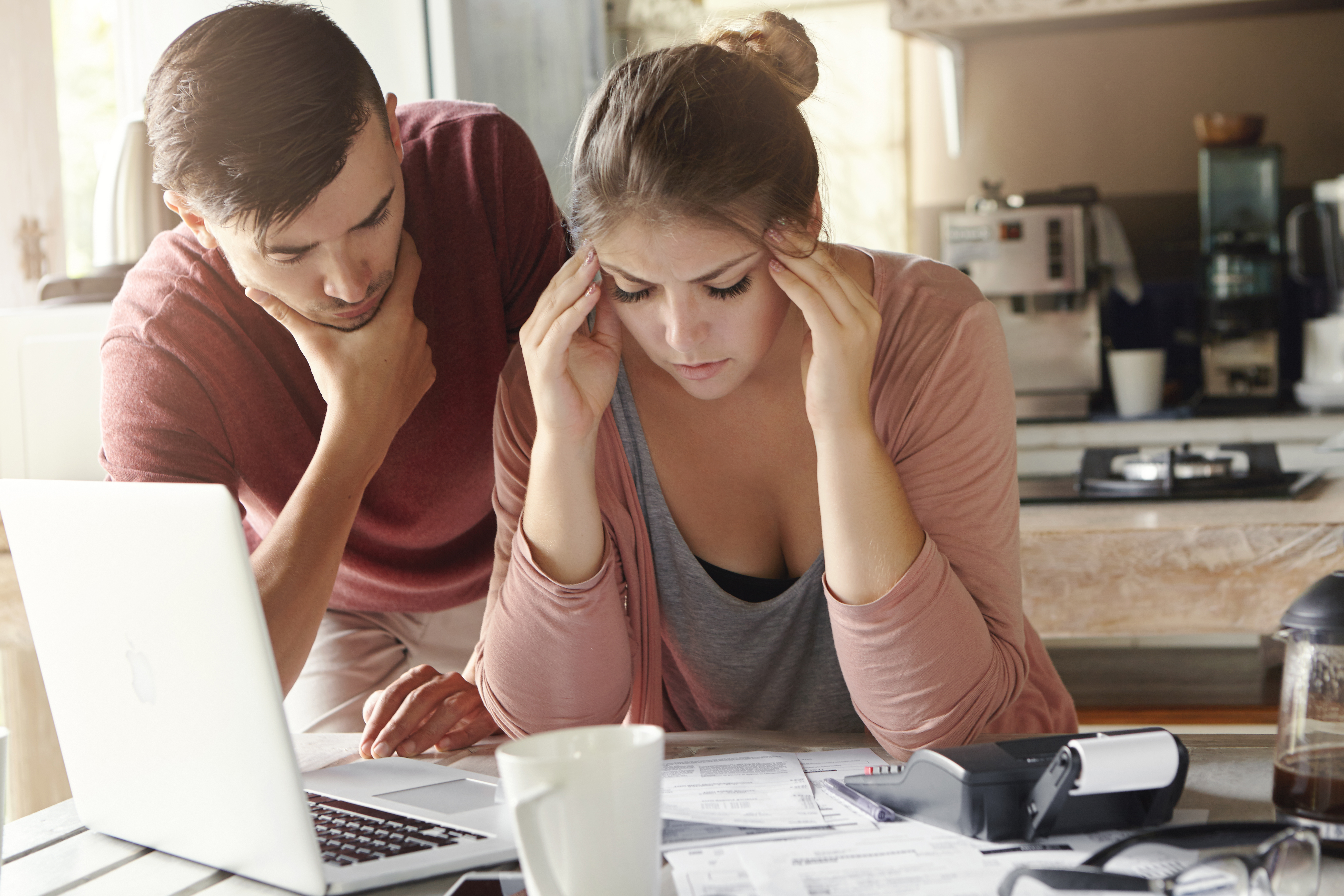 Young family with many debts facing financial stress. Unemployed male looking pensive standing next to his depressed wife with headache who is calculating expenses, trying to make both ends meet
